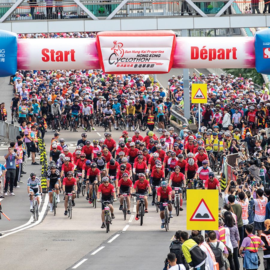 People riding bicycles on the road for the Hong Kong Cyclothon, passing the starting line inflatable arch while spectators look on.  