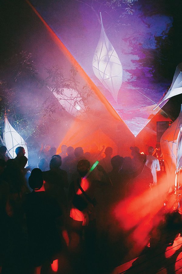 A crowd of people dancing under trees, surrounded by purple and red lighting.