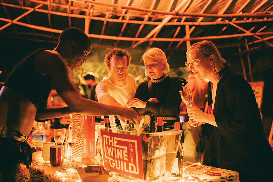 A group of people standing around a box of wine with a label on it that says “The Wine Guild”. 