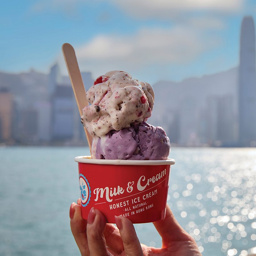 A hand holding two scoops of ice cream in a red “Milk & Cream” cup against Hong Kong’s daytime skyline. 