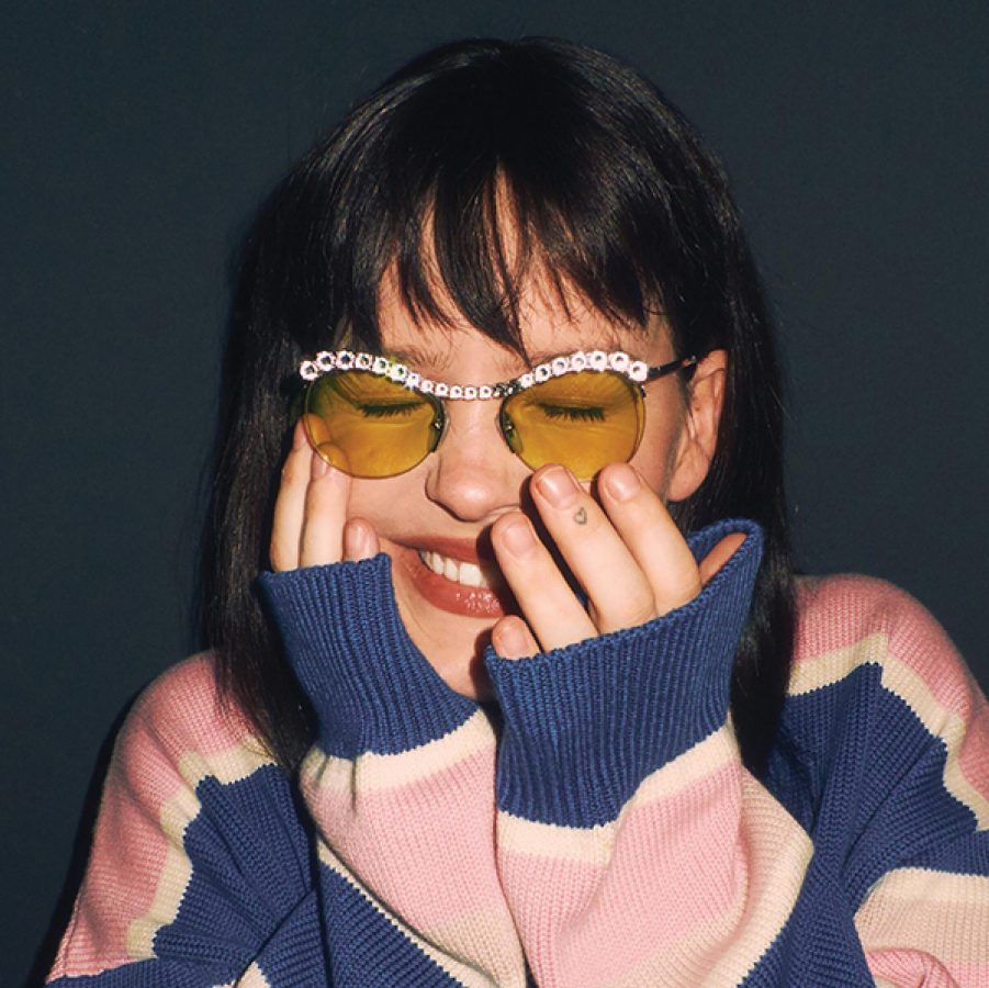 Singer Anne-Marie smiling with her hands on her face, wearing a striped pink-and-blue sweater and sunglasses.
