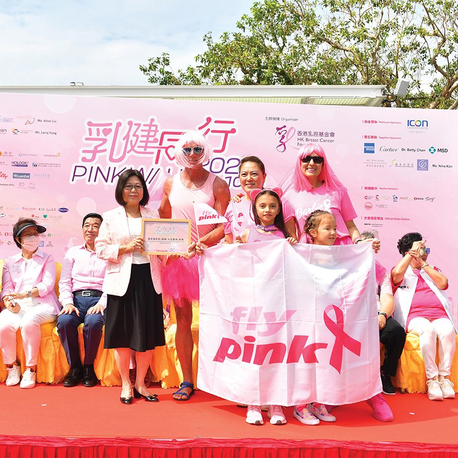 A group of people dressed in pink standing against a Pink Walk backdrop and holding a banner with a pink breast cancer ribbon and “fly pink” text on it.