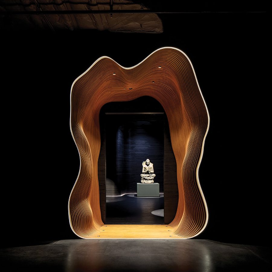 A sculpture displayed in a dark room, as seen through a lit-up, geometric arch. 