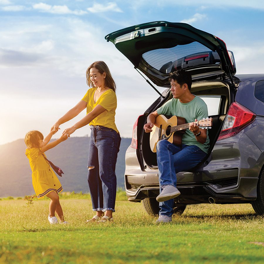A family of three spending time outdoors next to a car, with the father sitting on the trunk strumming a guitar while the mother and her daughter are playing together.