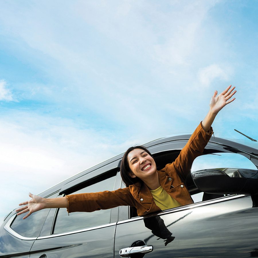 A smiling woman leaning out of a car window with her arms oustretched and a blue sky in the background.