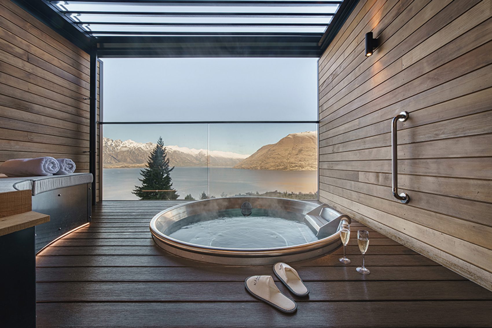 A hot tub inside a wood-panelled room, with a large window looking out onto a lake and mountains 