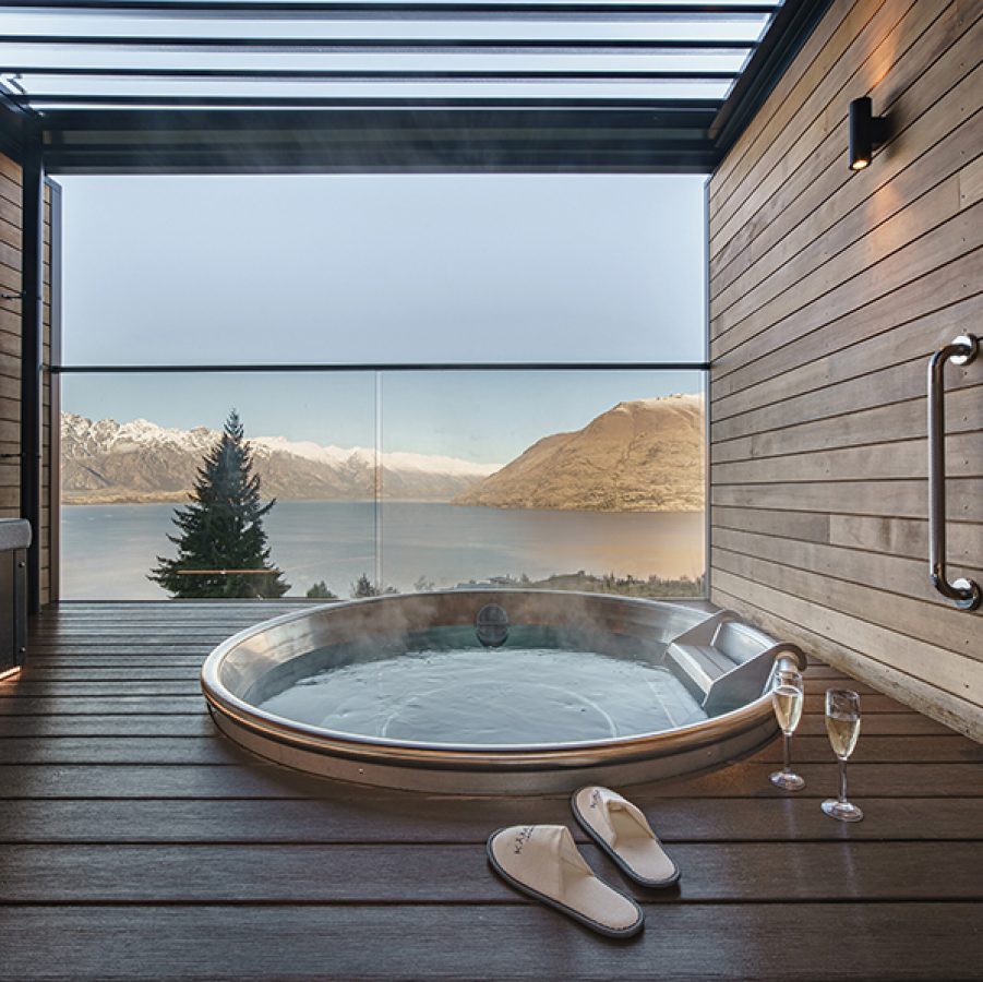 A hot tub inside a wood-panelled room, with a large window looking out onto a lake and mountains 