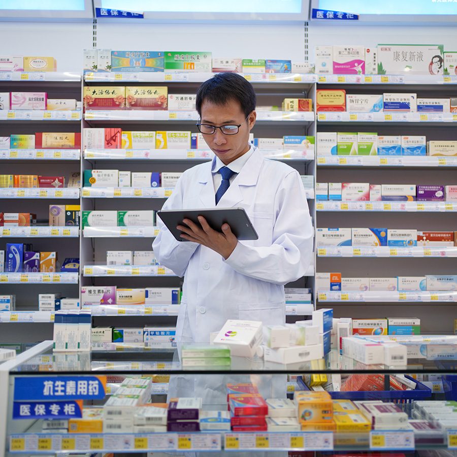 A man in a white lab coat stands at a pharmacy counter surrounded by boxes of medication. 