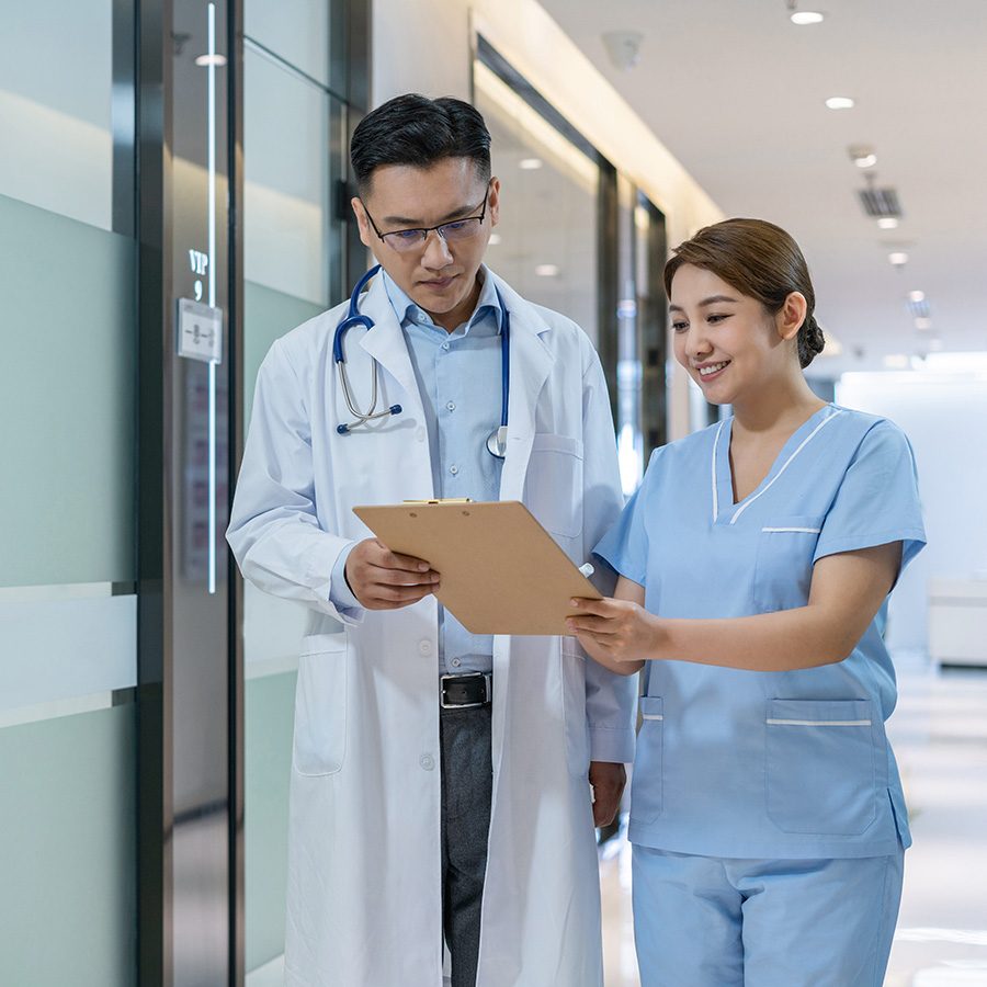 A male doctor in a white lab coat with a stethoscope around his neck shows a chart to a female health worker in blue scrubs. 
