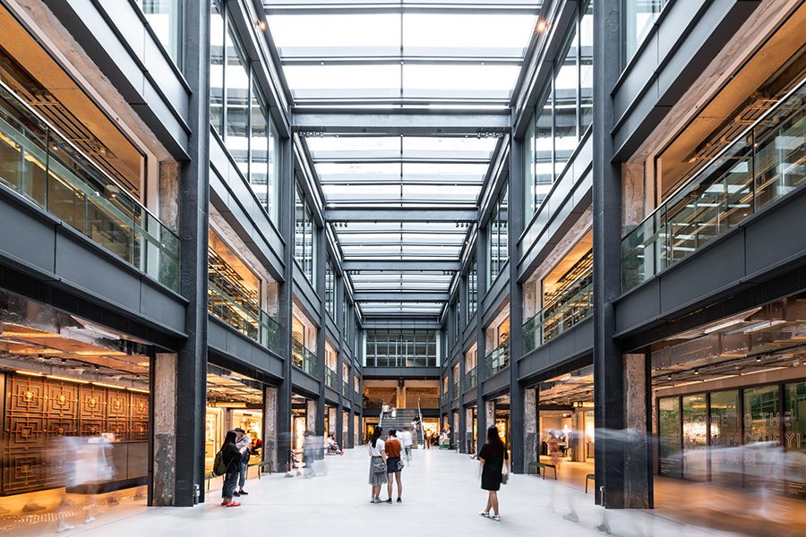 People walking inside The Mills. Its interior includes a glass ceiling, glass link bridges and steel columns. 