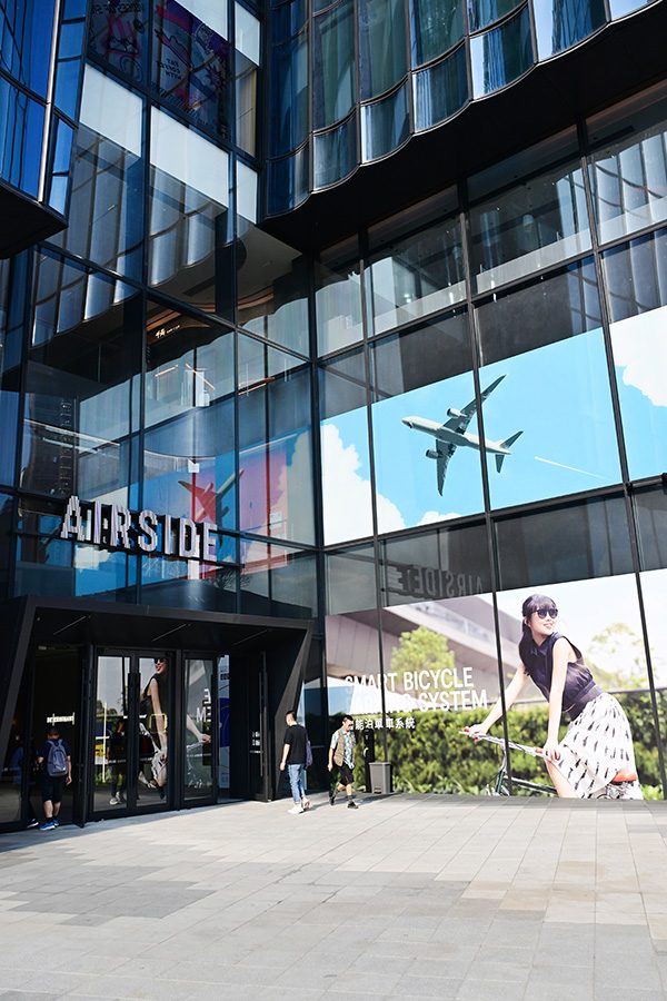 People walking in front of Airside. Its façade is composed of curved fluting glass, through which you can see images of an airplane in the sky and a woman on a bike.  