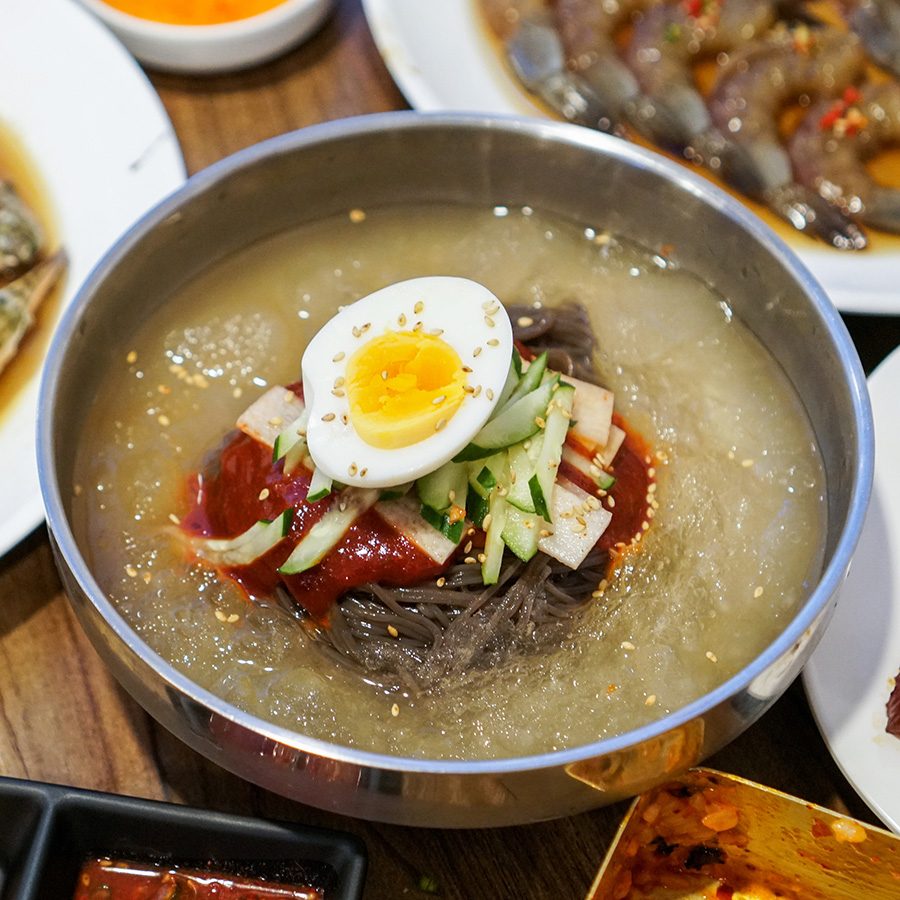 A steel bowl of naengmyeon cold buckwheat noodles topped with sliced vegetables and a sliced egg, served on a dining table full of various Korean dishes 