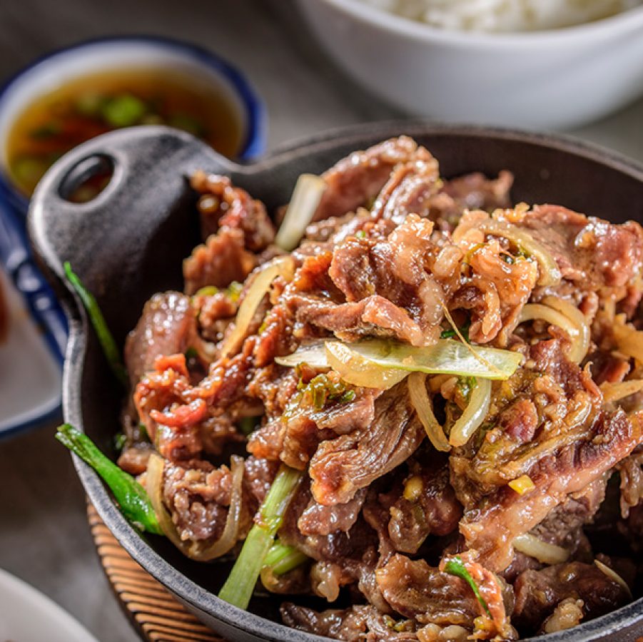 A closeup of a hot plate of beef bulgogi on a dining table laden with various Korean food