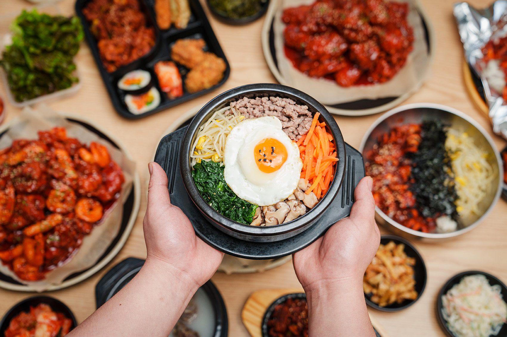  A closeup of a person holding a bowl of bibimbap over a dining table laden with various Korean dishes