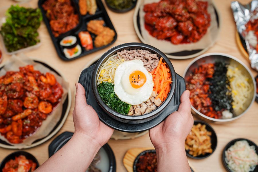  A closeup of a person holding a bowl of bibimbap over a dining table laden with various Korean dishes