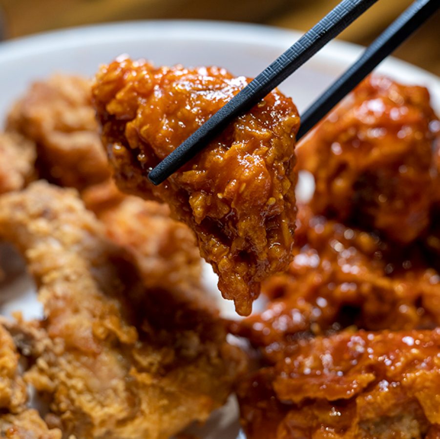 A closeup of chopsticks picking up a piece of maekdom-dak gangjeong from a plate of Korean sweet and sour fried chicken 