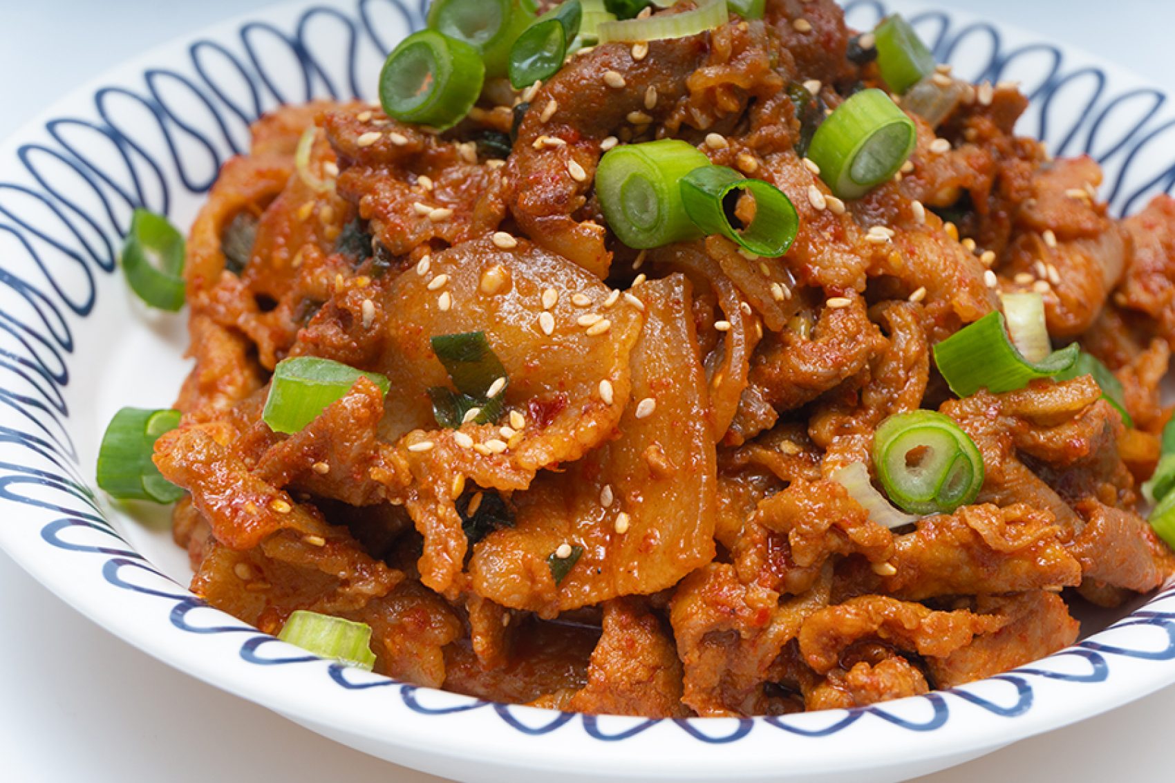 A closeup of a white and blue plate of jaeyook beokkeum, Korean spicy stir-fried pork, garnished with sesame seeds and chopped scallion spring onions
