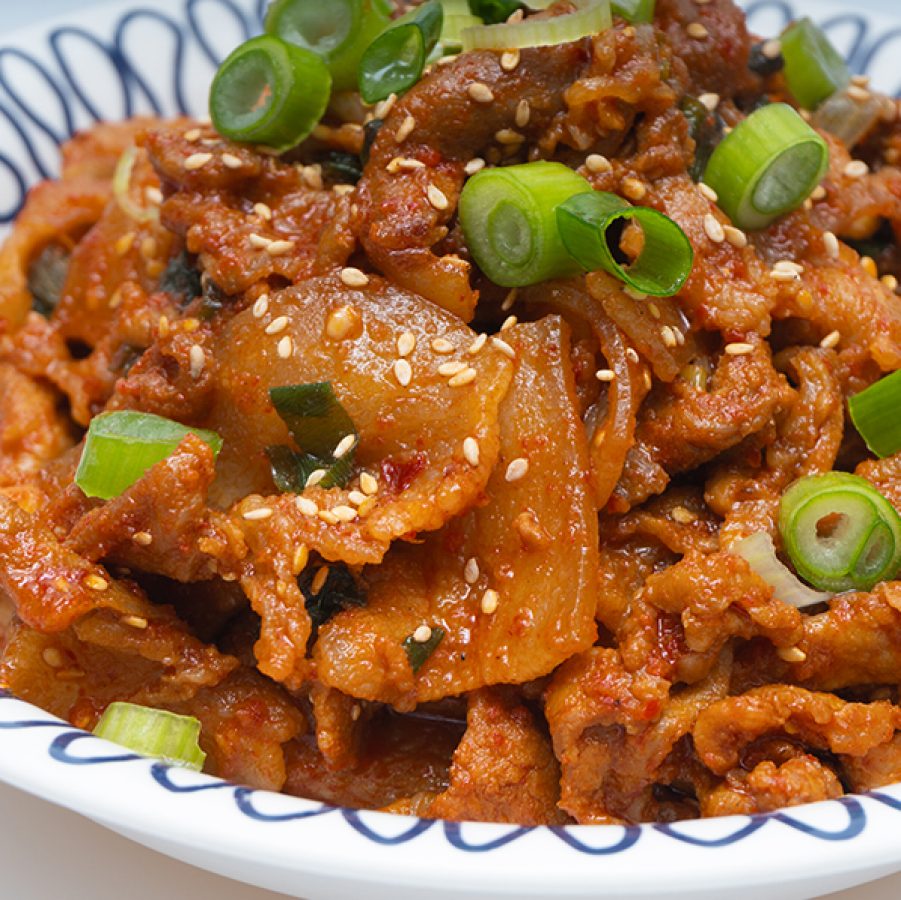 A closeup of a white and blue plate of jaeyook beokkeum, Korean spicy stir-fried pork, garnished with sesame seeds and chopped scallion spring onions