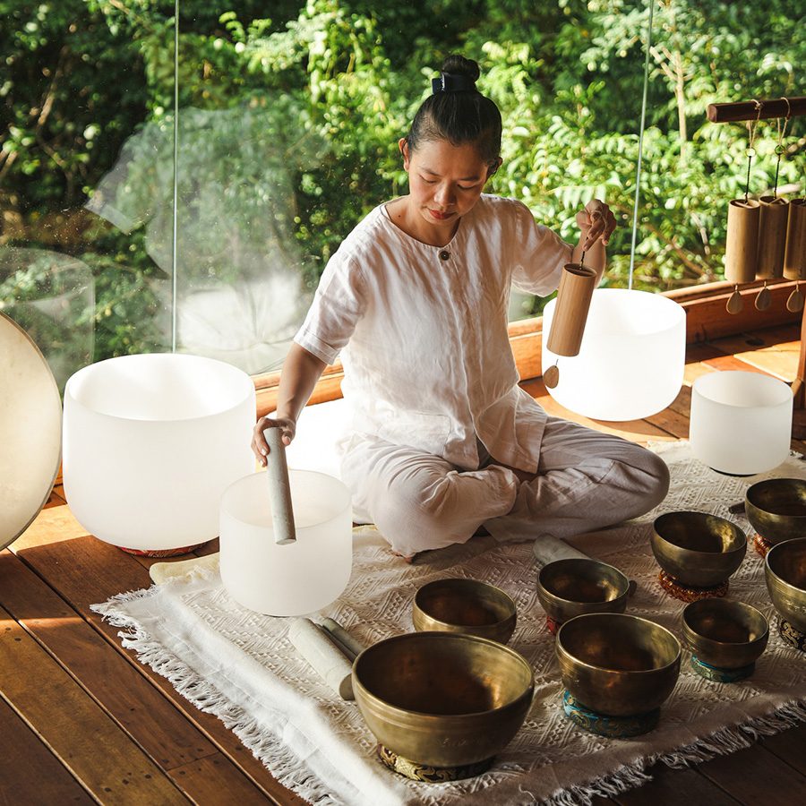 A woman sits on the floor playing singing bowls in front of a large window with a view of nature in the backdrop during daytime at Six Senses Ninh Van Bay spa in Vietnam 