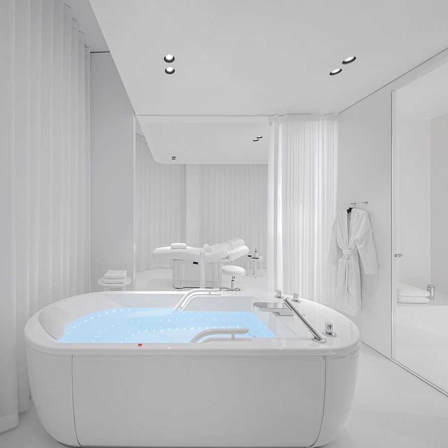 A tub in a room with white walls and curtains at Lanserhof Sylt. A bathrobe hangs on a wall on the right and a white recliner is in the background. 