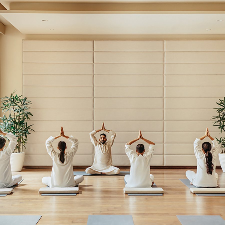  A group of four people in white kurtas sit cross-legged, facing their yoga instructor at Six Senses Vana. They are all holding their hands above their heads in a yoga pose. 