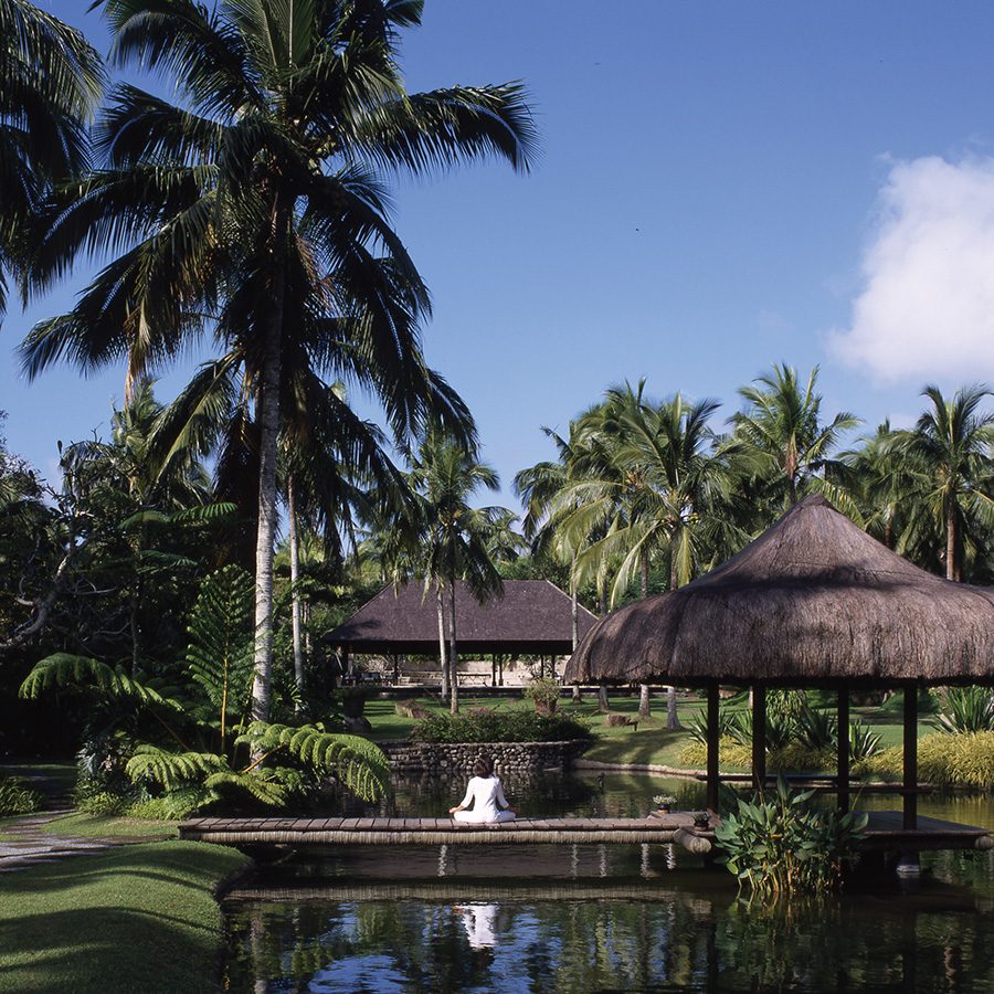 A woman in white sits cross-legged in a meditation pose on a bridge over a lagoon at The Farm at San Benito.