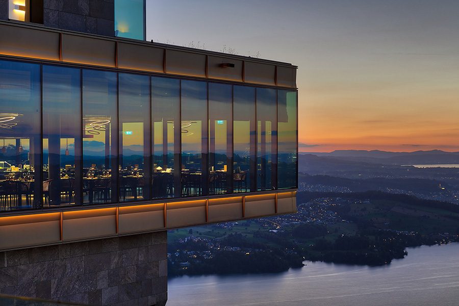 The dining room at Bürgenstock Hotel & Alpine Spa, which features floor-to-ceiling windows all the way around, is seen from the outside against a sunset backdrop