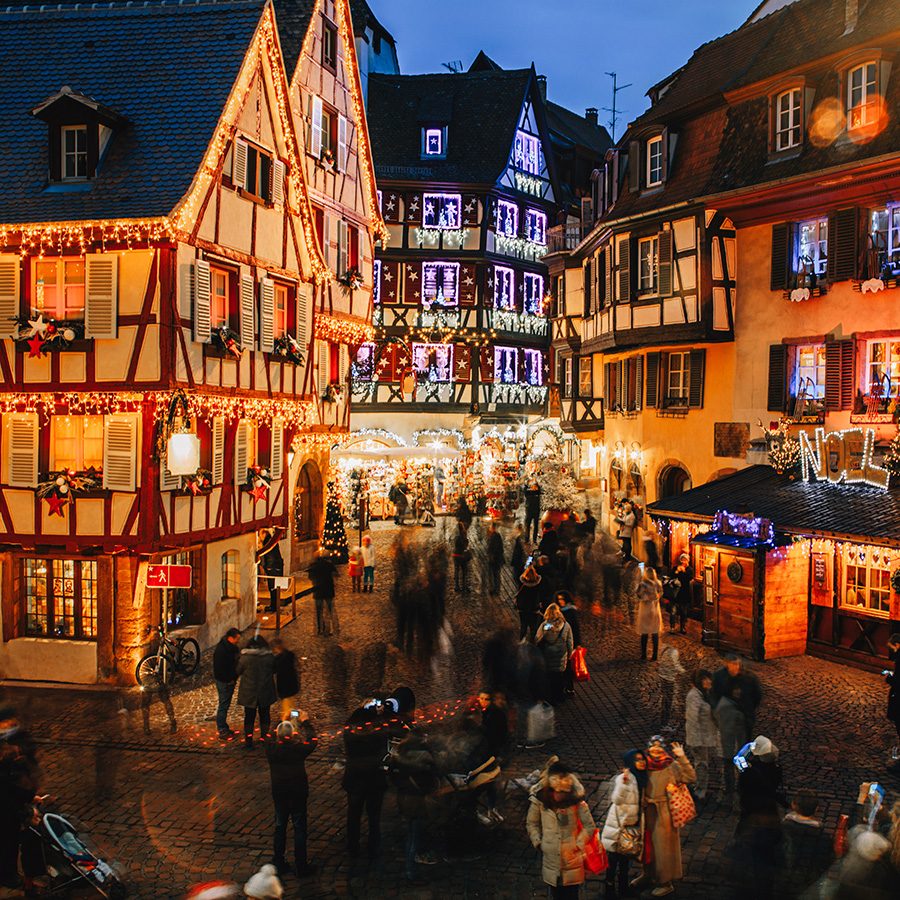 Crowds wander among traditional timber-frame houses illuminated by Christmas lights at night in Colmar.  