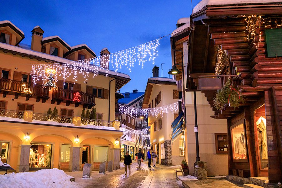 People walk down a narrow, snow-covered street lined with traditional alpine buildings, as Christmas lights hang overhead, in Aosta. 