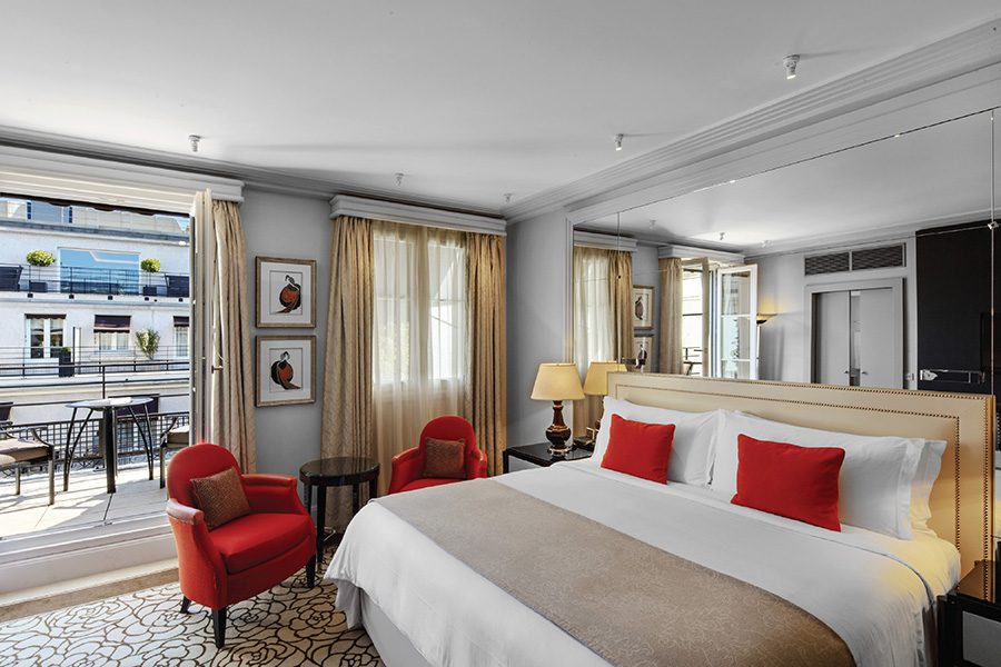 A bright guestroom with a large bed, two red armchairs and a set of French windows leading to a sunlit terrace, at Prince de Galles, a Luxury Collection Hotel 