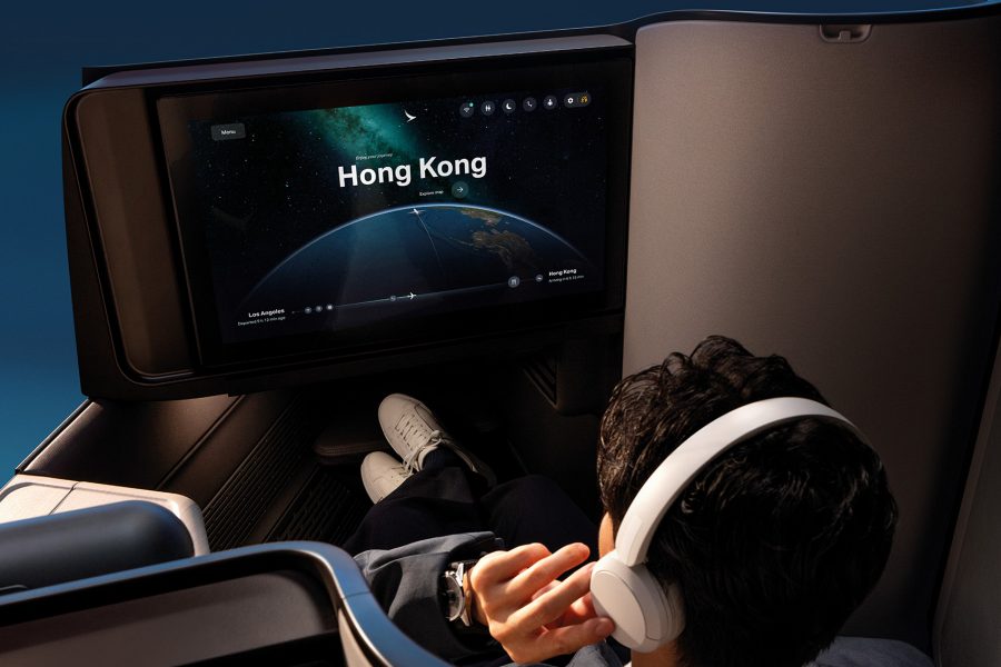 A man with white headphones and white sneakers, sitting in Cathay Pacific’s new Aria Suite. The screen in front of him displays the route from Los Angeles to Hong Kong.