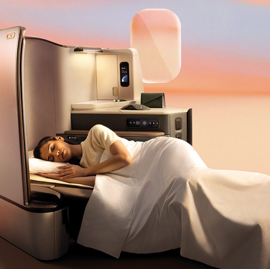A woman covered by a blanket, sleeping on the fully lie-flat bed in Cathay Pacific’s new Aria Suite. There is a sunset-coloured gradient background behind her.