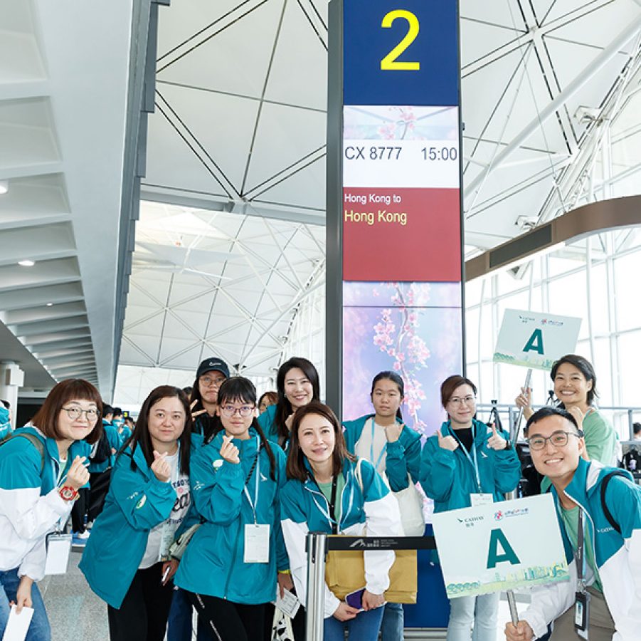 A group shot of people dressed in Cathay Pacific jackets standing by an airport boarding gate sign.