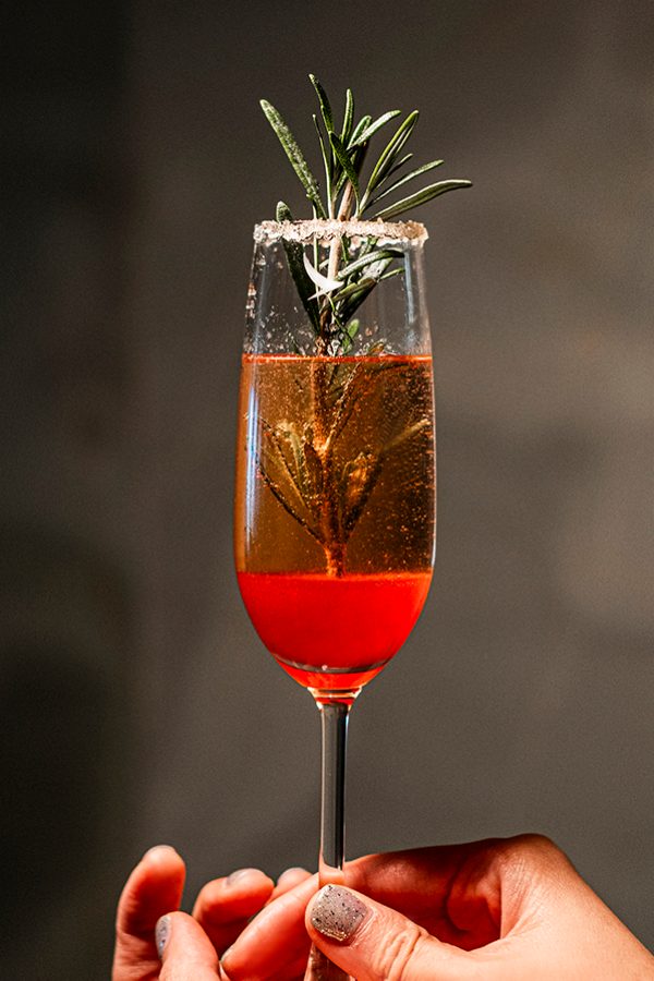 Two hands holding a raspberry champagne cocktail in a champagne flute, featuring a sprig of rosemary. 