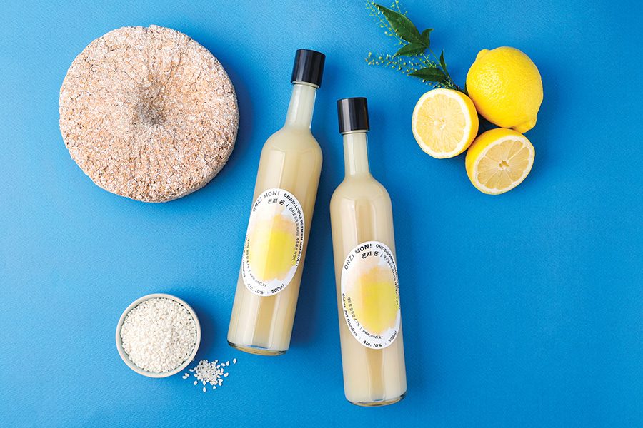 Bottles of makgeolli from Onzi Brewery placed against a blue background next to bowls of rice and lemons.