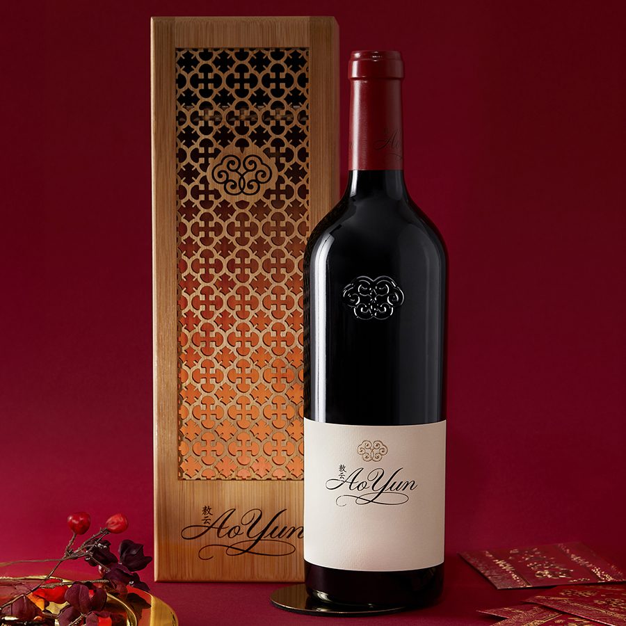 A bottle of Ao Yun wine next to its decorative box against a red backdrop. Red envelopes are scattered next to the bottle.