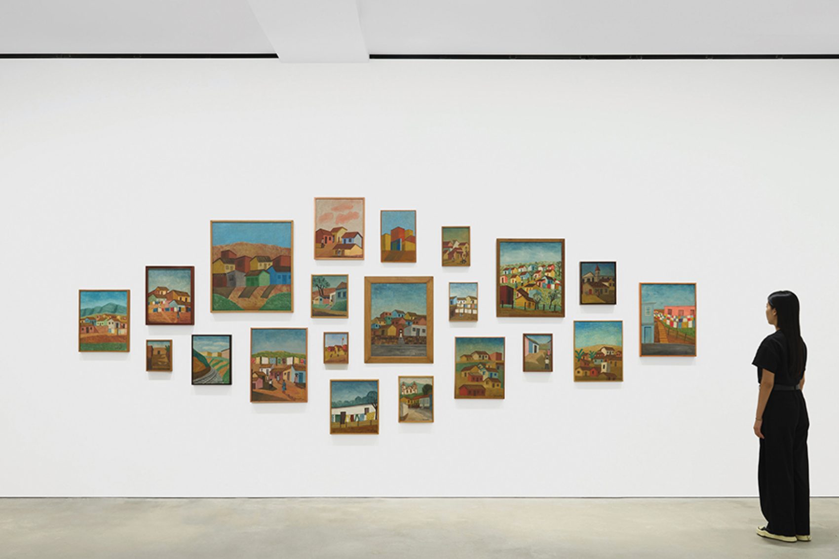 A woman in black clothes stands to the right, looking at a group of Amadeo Luciano Lorenzato’s paintings on the wall. 