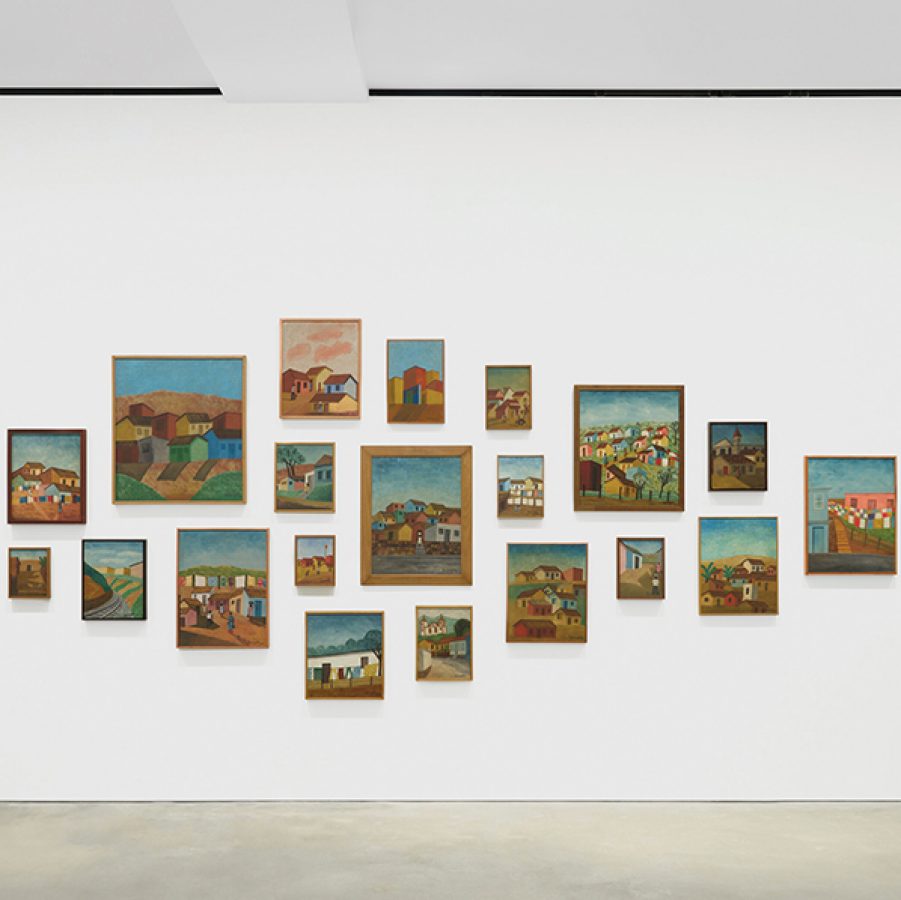 A woman in black clothes stands to the right, looking at a group of Amadeo Luciano Lorenzato’s paintings on the wall. 