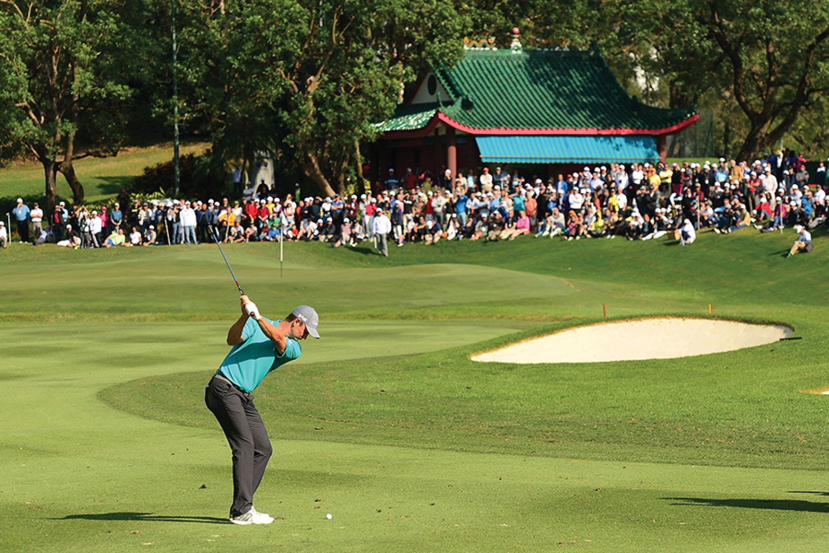 A man swinging a club at Hong Kong Golf Club in Fanling, while a large crowd in front of a green pavilion watches on. 