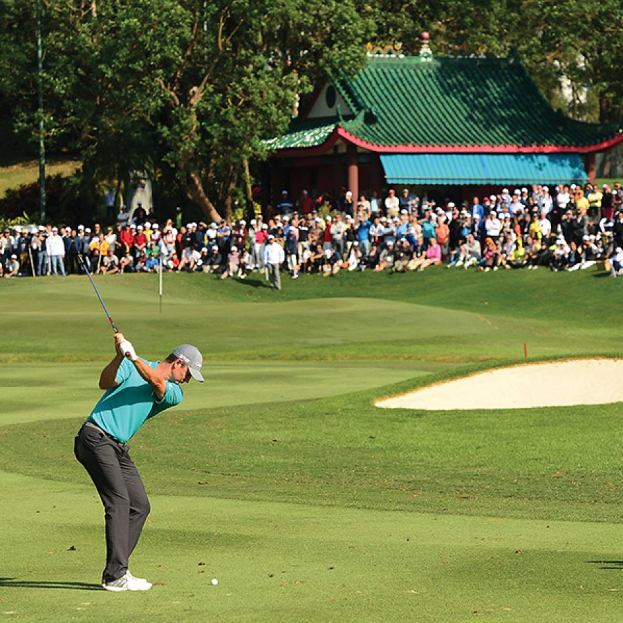 A man swinging a club at Hong Kong Golf Club in Fanling, while a large crowd in front of a green pavilion watches on. 