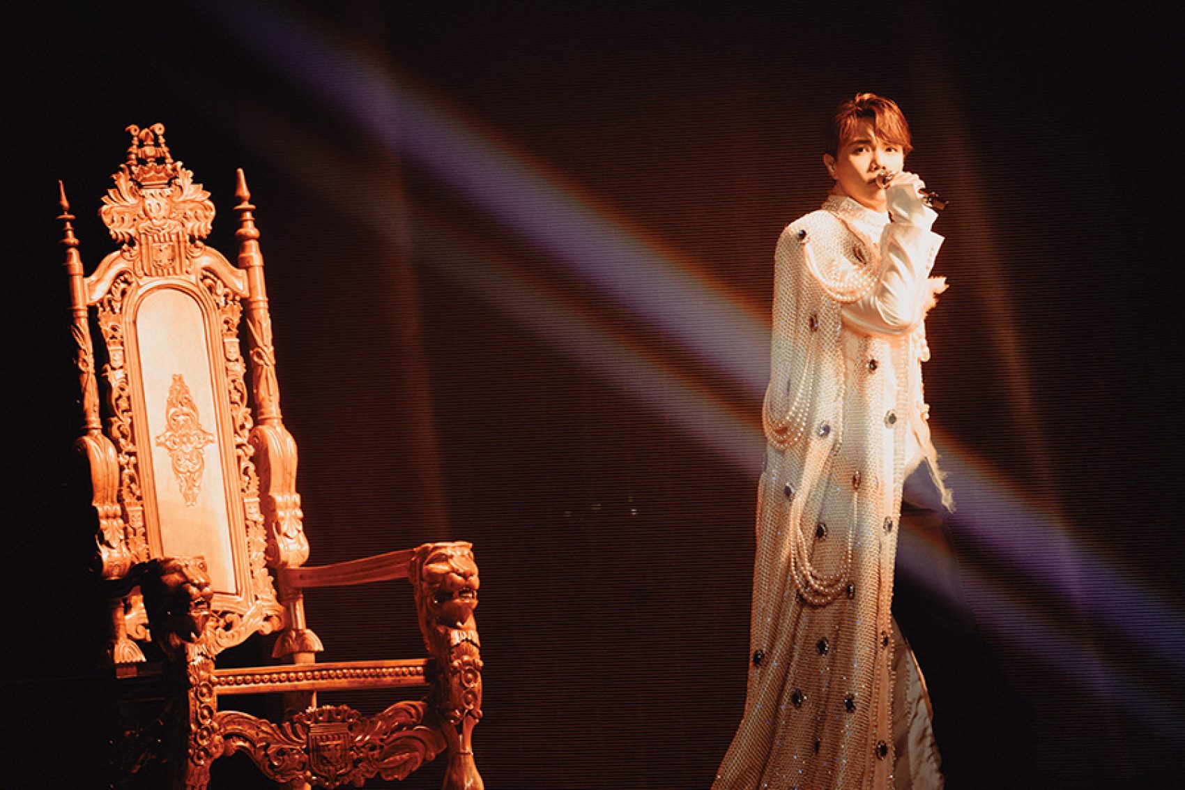 Hins Cheung on stage in a sparkly, floor-length robe, standing in front of a throne and holding a microphone. 