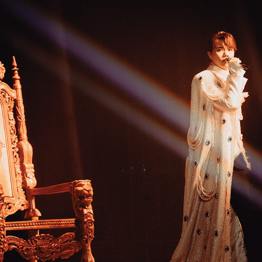 Hins Cheung on stage in a sparkly, floor-length robe, standing in front of a throne and holding a microphone. 