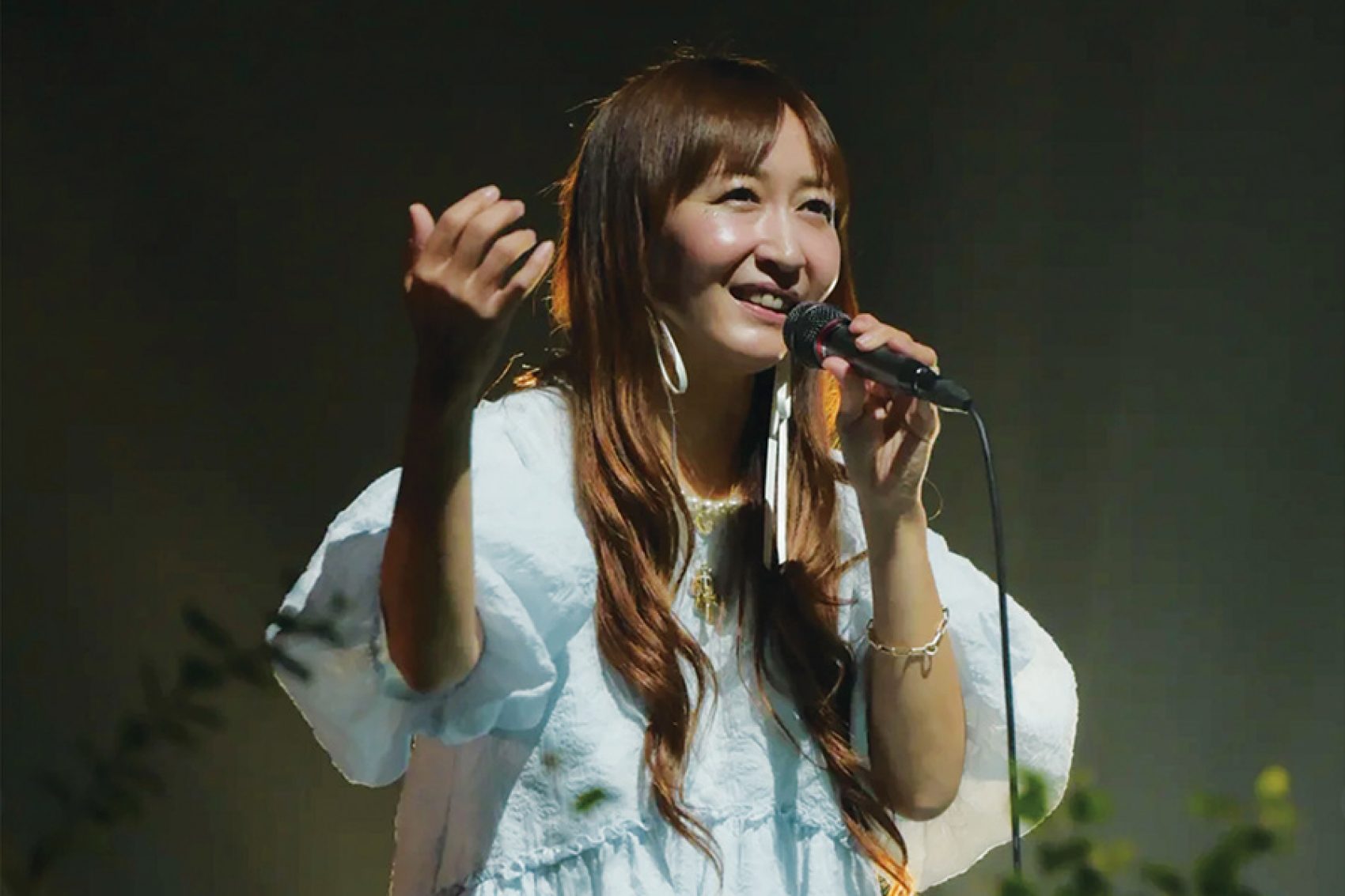 Kokia holding a microphone on stage, dressed in white. 