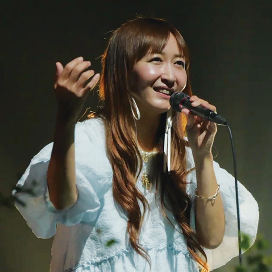 Kokia holding a microphone on stage, dressed in white. 