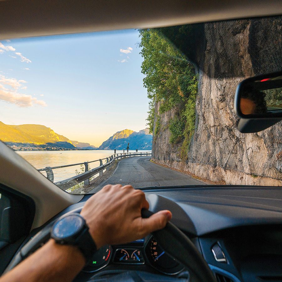 A person driving a car with a hand on the wheel, with a view of a winding road next to a cliff