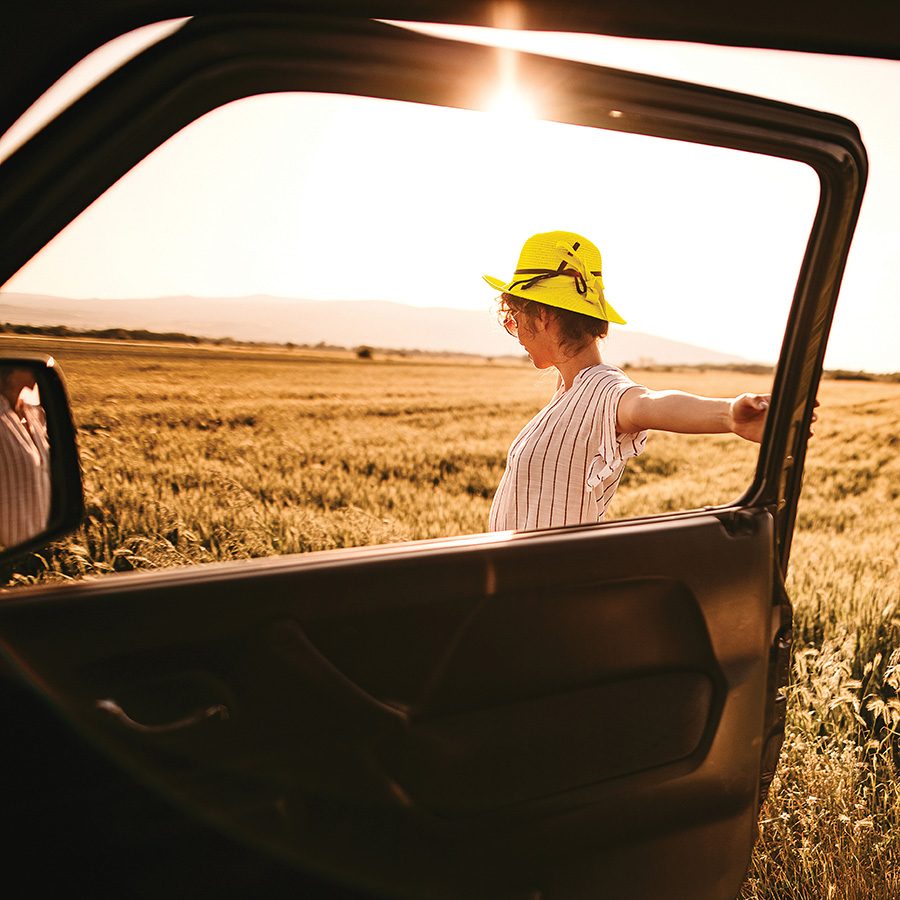 A woman holds the door of a car open in a large field of grass