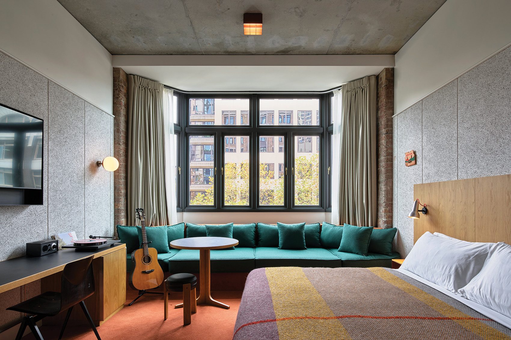 A guestroom at Ace Hotel Sydney featuring a bed with a striped blanket, a minimalist-style desk and coffee table, and a long green sofa below a large window. Accessories include a guitar, radio and record player. 