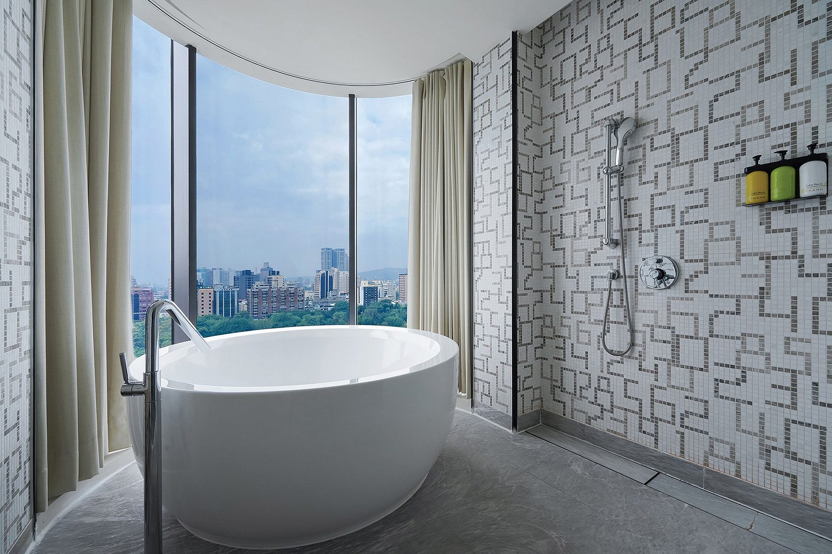 A spacious, modern bathroom at Hotel Indigo Kaohsiung Central Park. A large freestanding bath is placed in front of a curved, floor-to-ceiling window that offers city views.