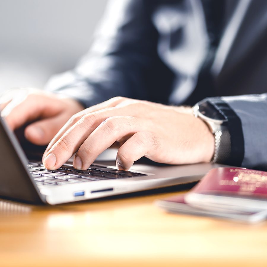 A man typing on a laptop with his passport and travel documents beside him. 
