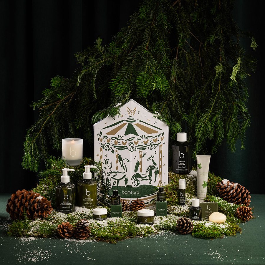 Skincare and self-care products from Bamford, available as gift sets this holiday season, arranged neatly among pine cones and draping coniferous tree branches. 